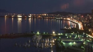 Un paisaje urbano frente al agua de noche, con edificios iluminados y un puente reflejado en el agua.