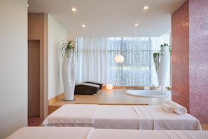 Un spa moderno con dos camillas de masaje, un amplio ventanal con cortinas translúcidas, una bañera redonda y un alto jarrón blanco con plantas.