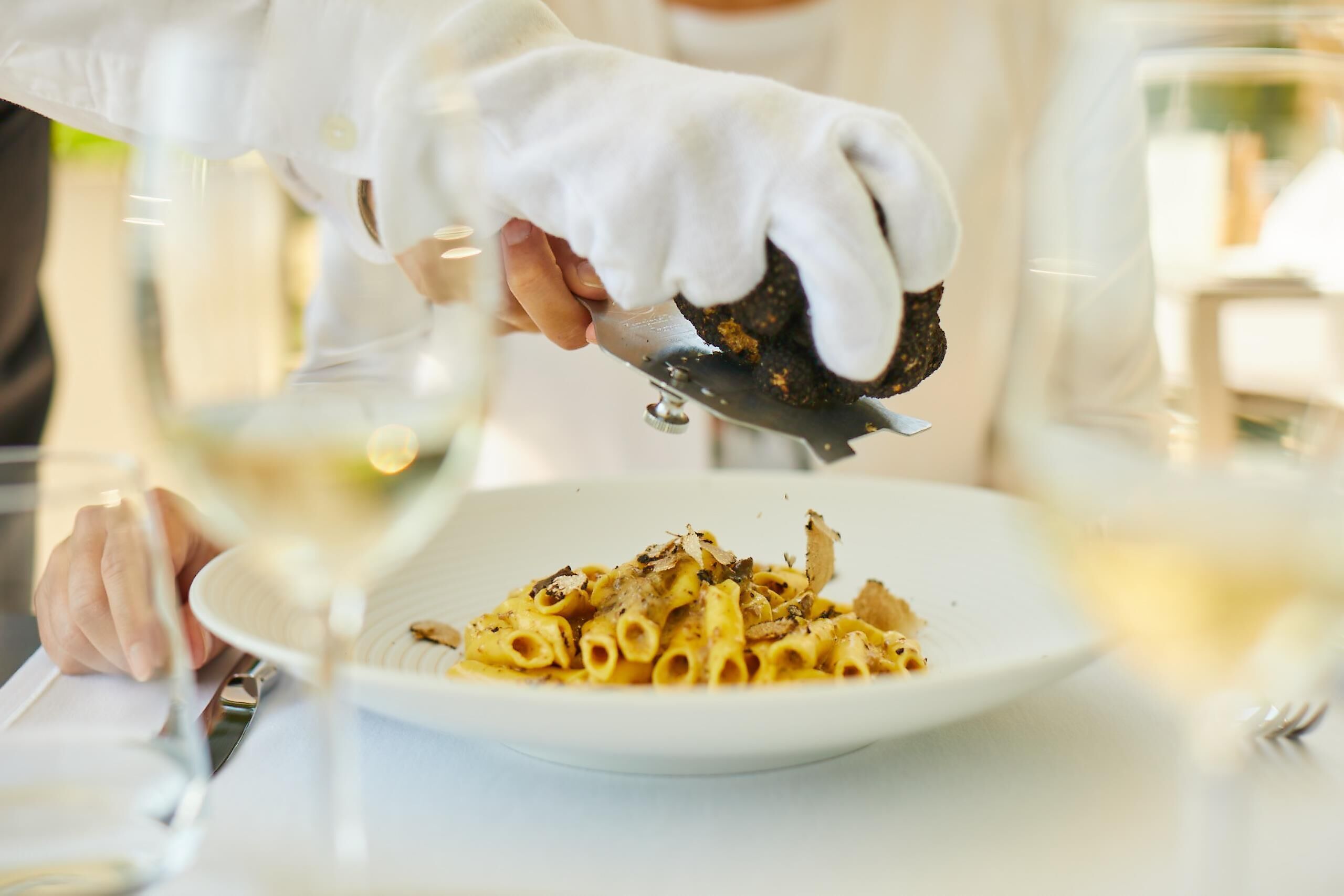 Una persona sirviendo pasta con un tenedor, un plato de pasta y una copa de vino blanco sobre una mesa.