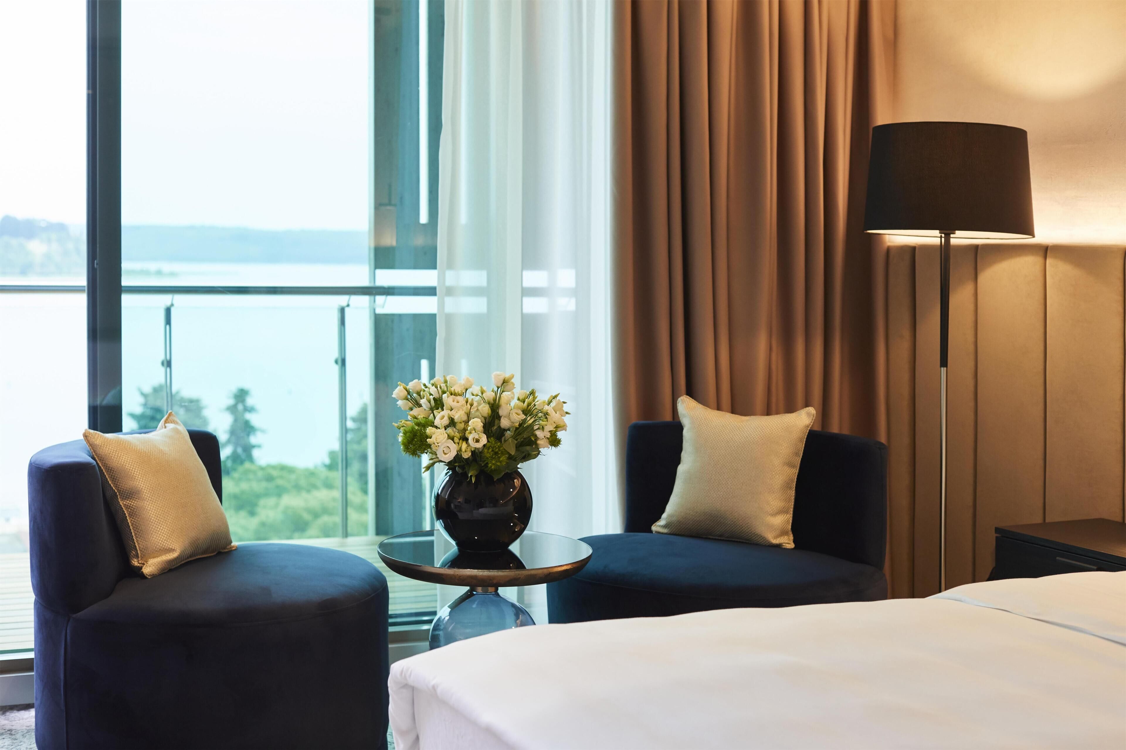 Ein Hotelzimmer mit einem Bett, zwei Sesseln, einem Tisch mit Blumenvase, einer Lampe und Meerblick.
