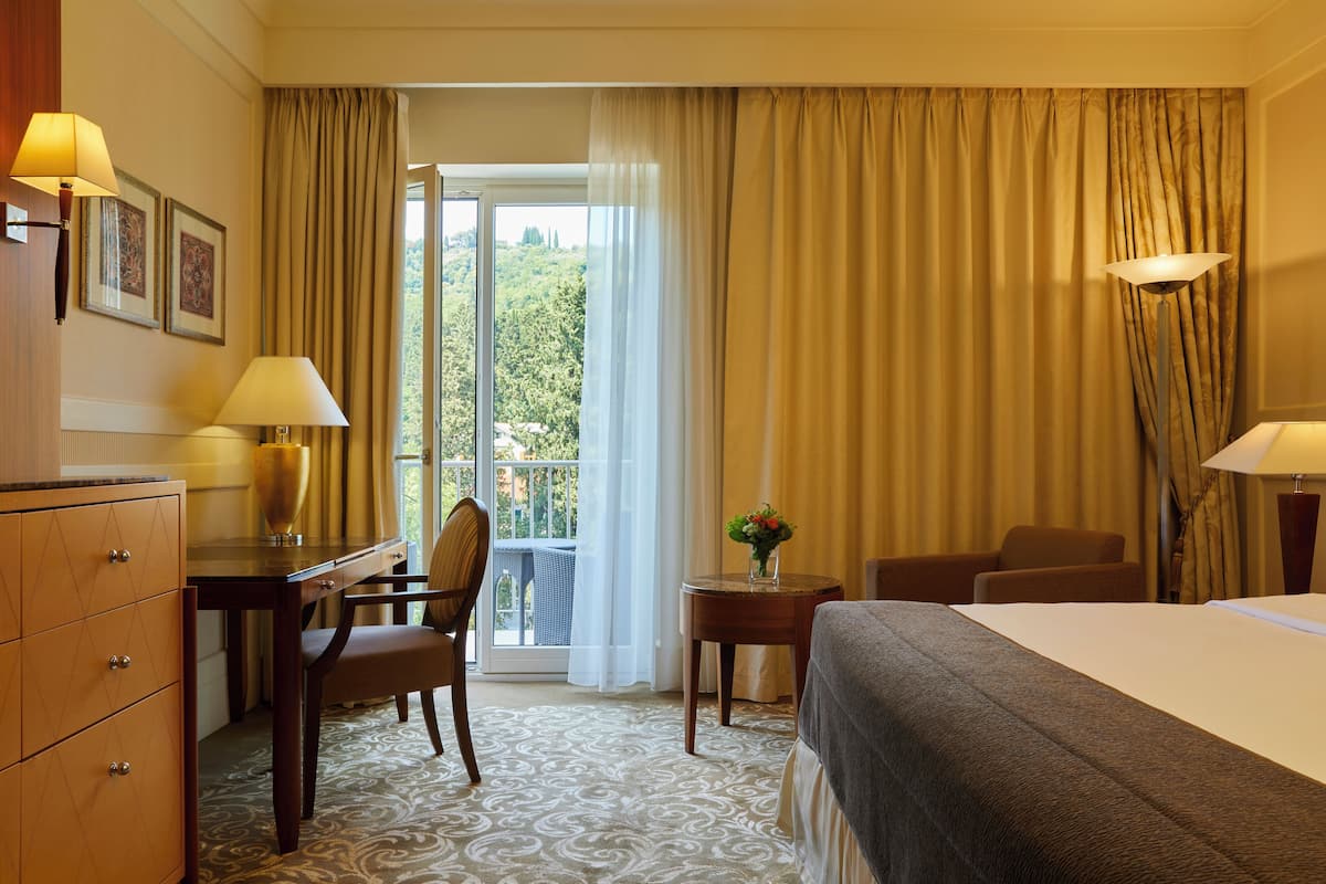 Ein Hotelzimmer mit Bett, Schreibtisch, Stuhl, einem kleinen Tisch mit Blumenvase und Blick ins Grüne durch das Fenster.