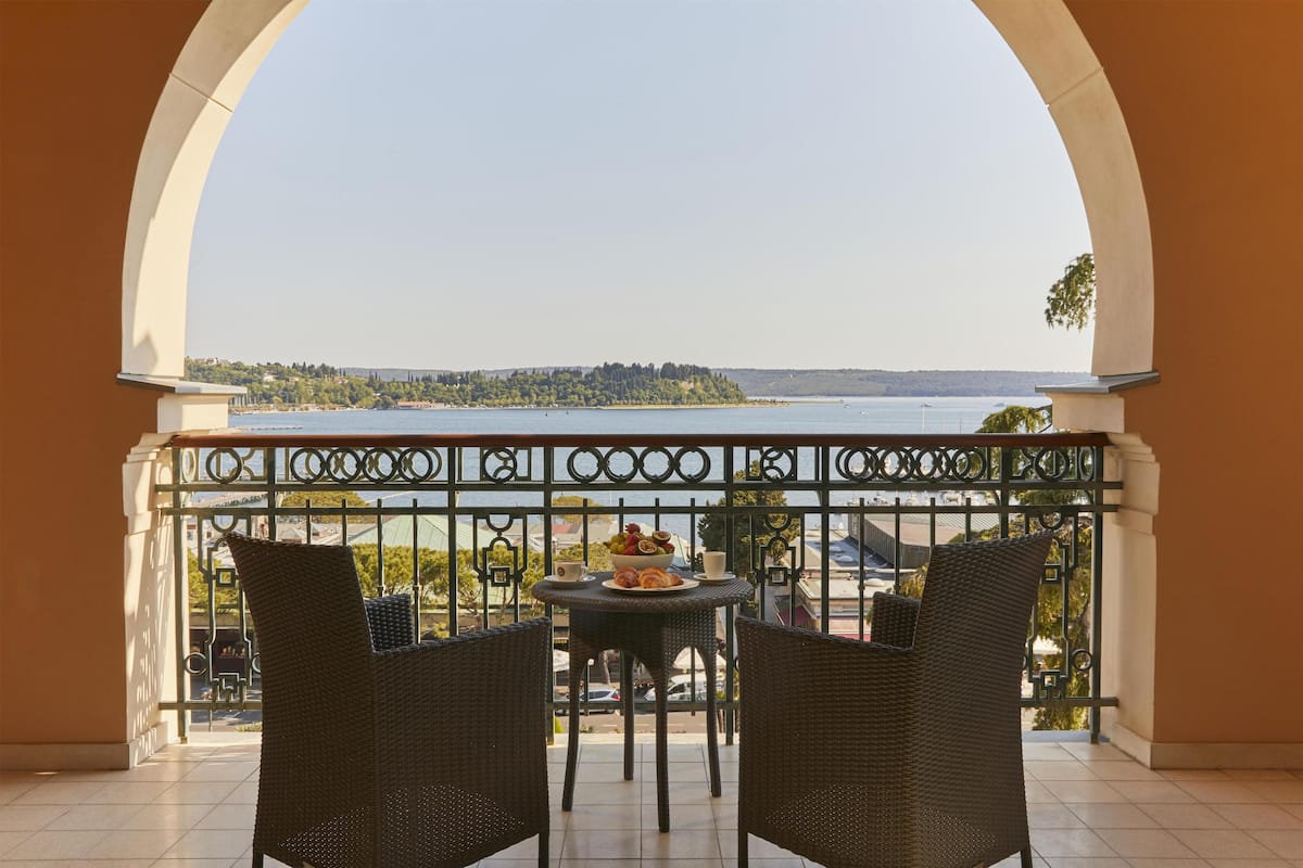 Ein Balkon mit Korbstühlen, einem Tisch mit Obstschale und Blick auf einen See sowie entfernte Bäume.