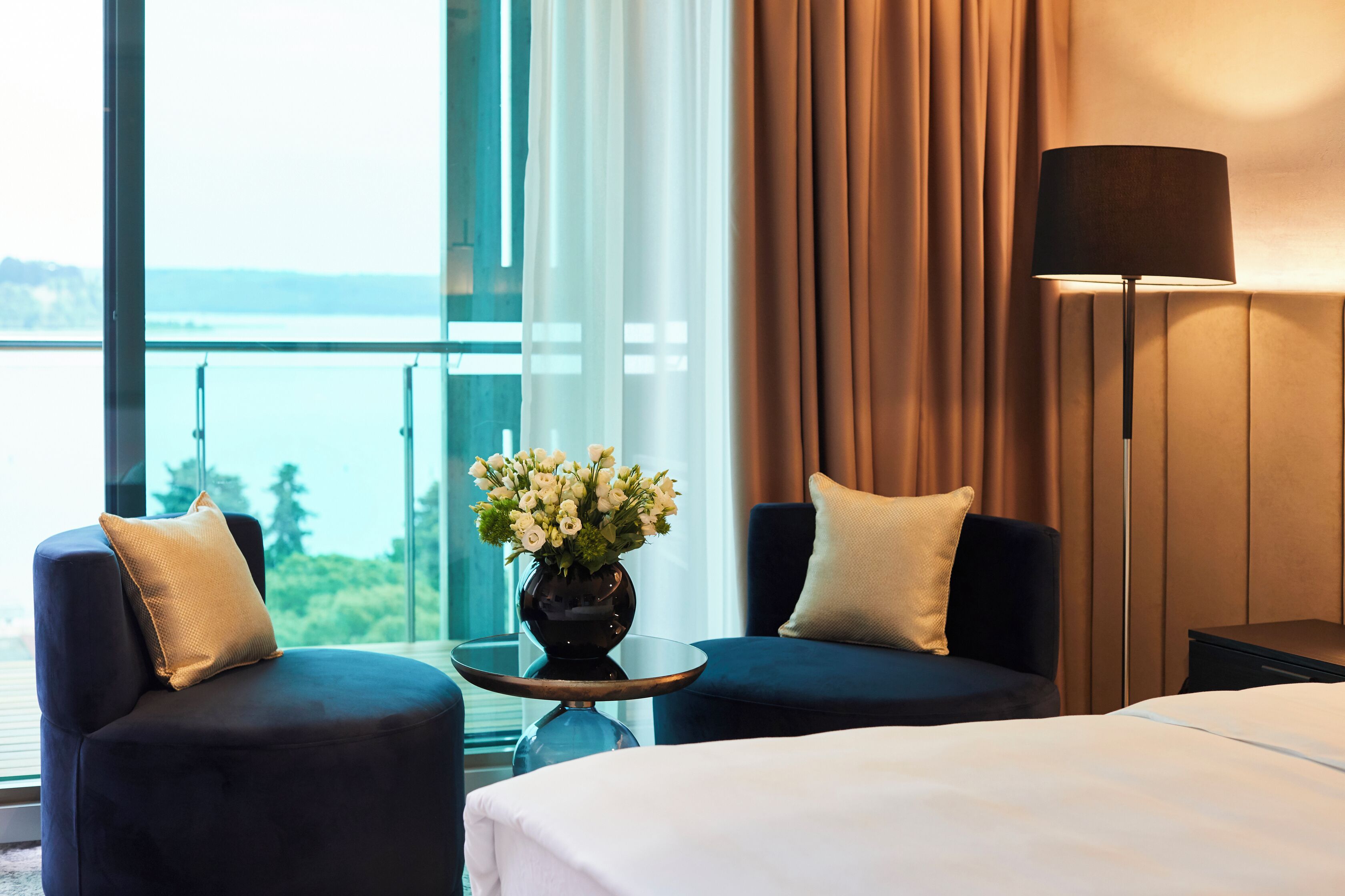 Une chambre d’hôtel avec un lit, deux fauteuils, une table ornée d’un vase de fleurs, une lampe et une vue sur la mer, visible à travers de grandes fenêtres.