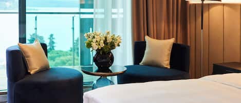 Une chambre d’hôtel avec un lit, deux fauteuils, une table ornée d’un vase de fleurs, une lampe et une vue sur la mer, visible à travers de grandes fenêtres.