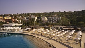 A beach with sun loungers and umbrellas, a pool, and buildings in the background. - Hotel Palace Portoroz Operated by Minor Hotels (Piran)