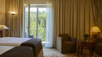 Une chambre d’hôtel avec un lit, deux chaises, une petite table sur laquelle se trouve un vase, et une vue sur une forêt, visible à travers une grande fenêtre.
