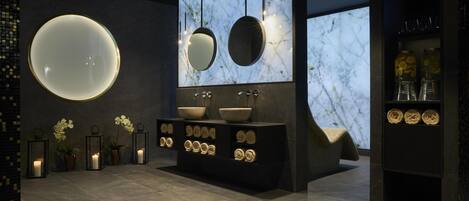 A modern bathroom with a large round mirror, two smaller mirrors, a central sink with a bowl, and a dark-toned vanity with decorative items.