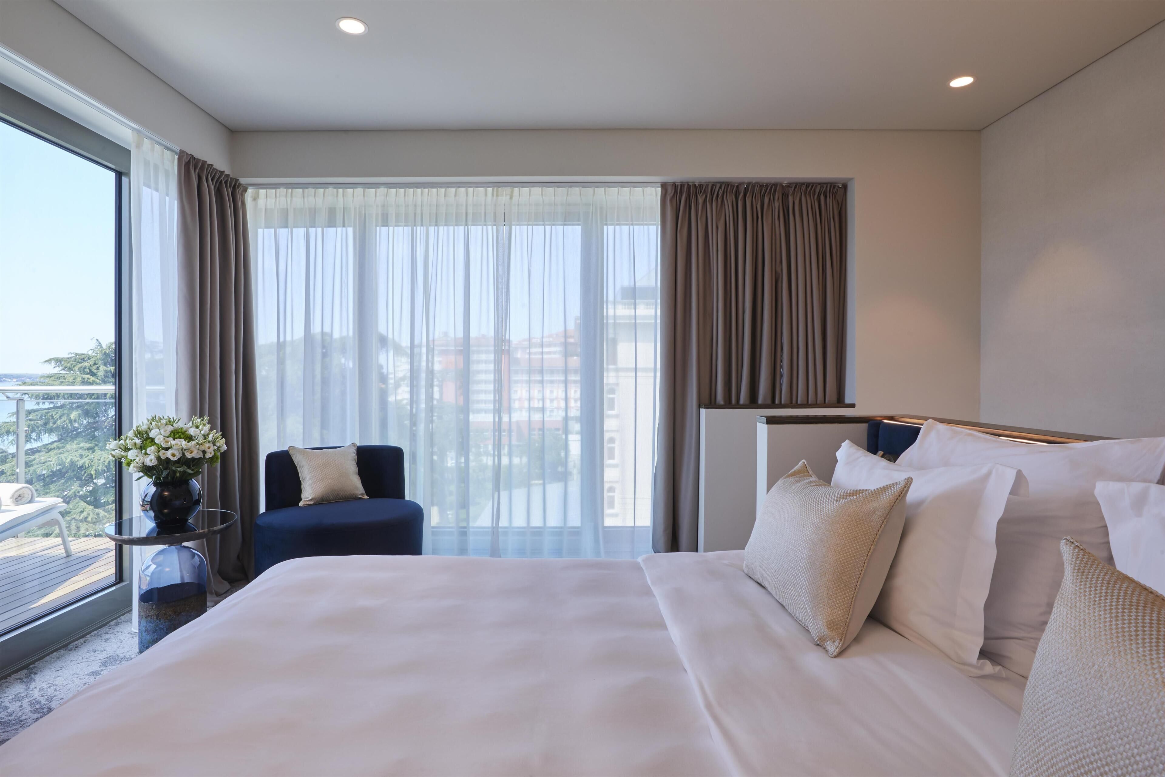 Un cuarto de hotel con una cama grande, una silla, una mesita con un florero y vistas de la ciudad a través de amplios ventanales.