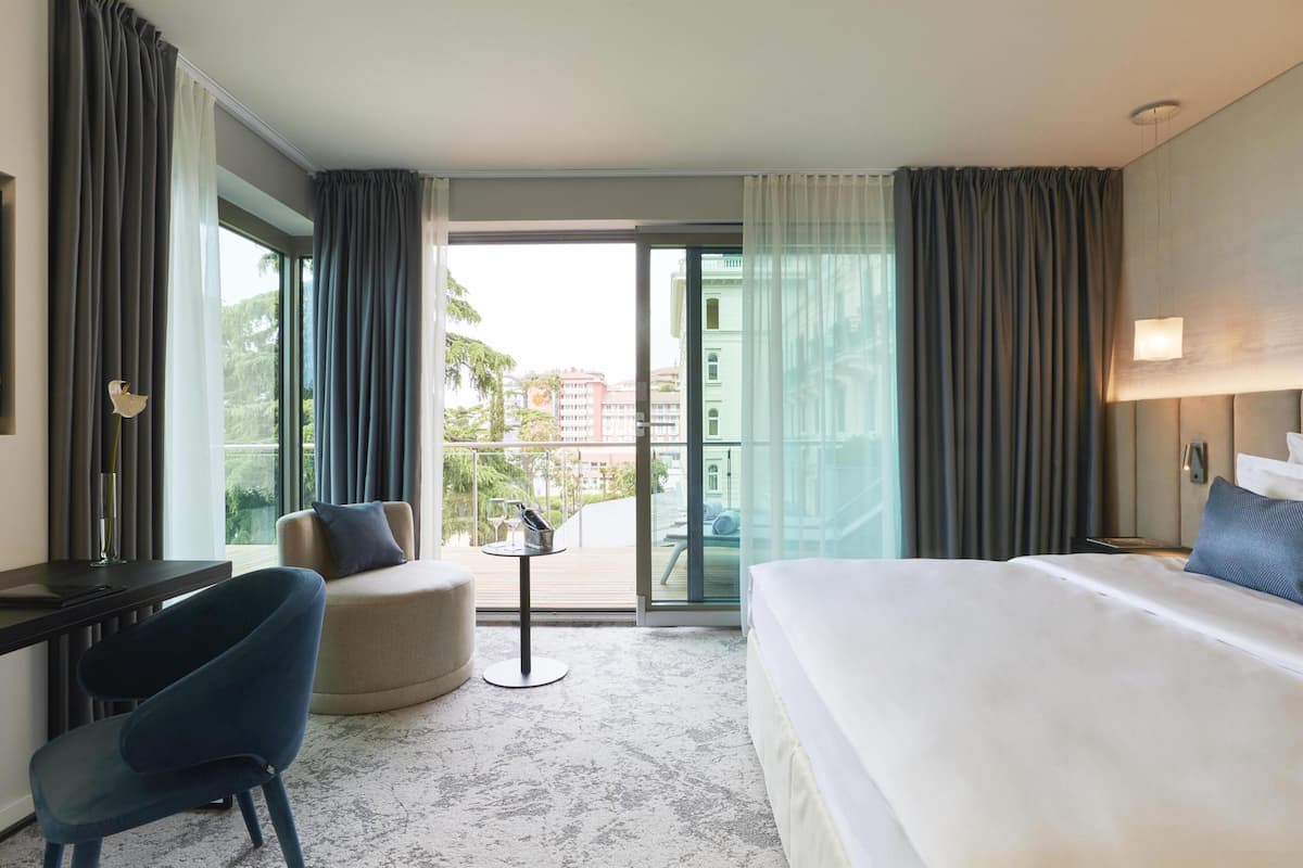 Ein Hotelzimmer mit Bett, Schreibtisch, Stuhl, einem kleinen Tisch und einem Balkon mit Blick auf Gebäude und Bäume.