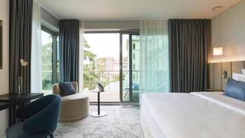 A hotel room with a bed, a desk, a chair, a small table, and a balcony with a view of buildings and trees.