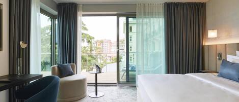 A hotel room with a bed, a desk, a chair, a small table, and a balcony with a view of buildings and trees.