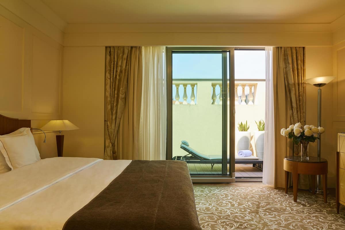 Ein Hotelzimmer mit Bett, Nachttisch mit Lampe, Blick auf einen Balkon mit Stuhl und Pflanzen sowie eine Vase mit Blumen auf einem Beistelltisch.