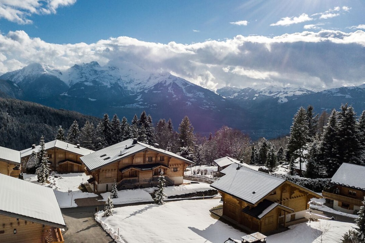 A cluster of wooden cabins with snow-covered roofs, set against a backdrop of snow-capped mountains and a clear sky.