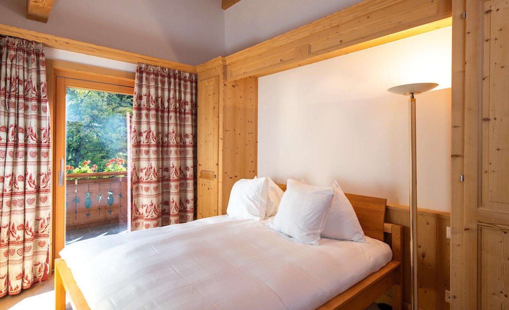 A bedroom with a wooden bed, white bedding, and a view of greenery through a window.