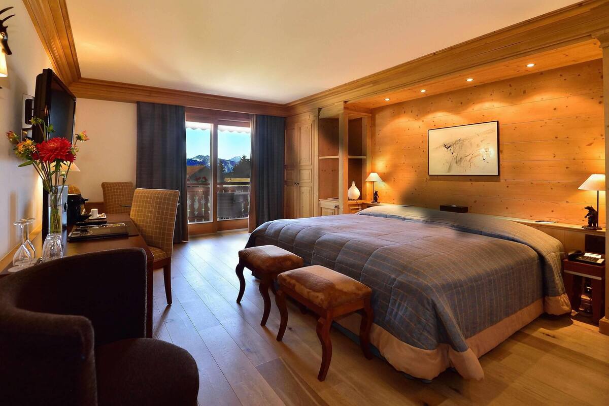 A spacious bedroom with a large bed, a wooden floor, a dining area with a table and chairs, and a balcony with a view.