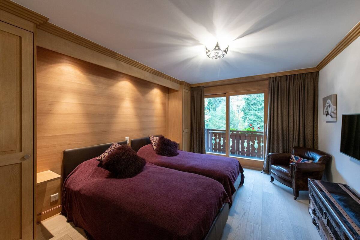 A room with two beds, a wooden wardrobe, a desk, a chair, and a large window with a view of trees.