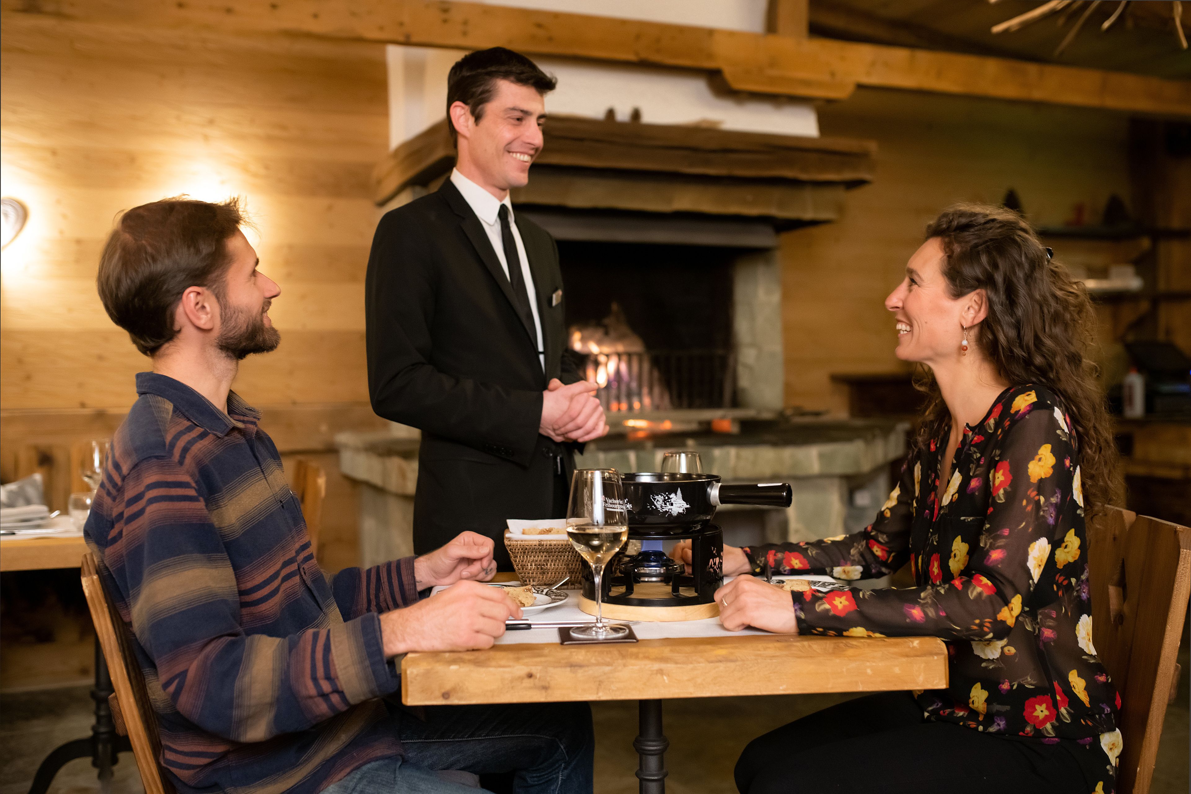 A wooden interior with a fireplace, a dining table with a hot plate, and a waiter serving a glass of wine.