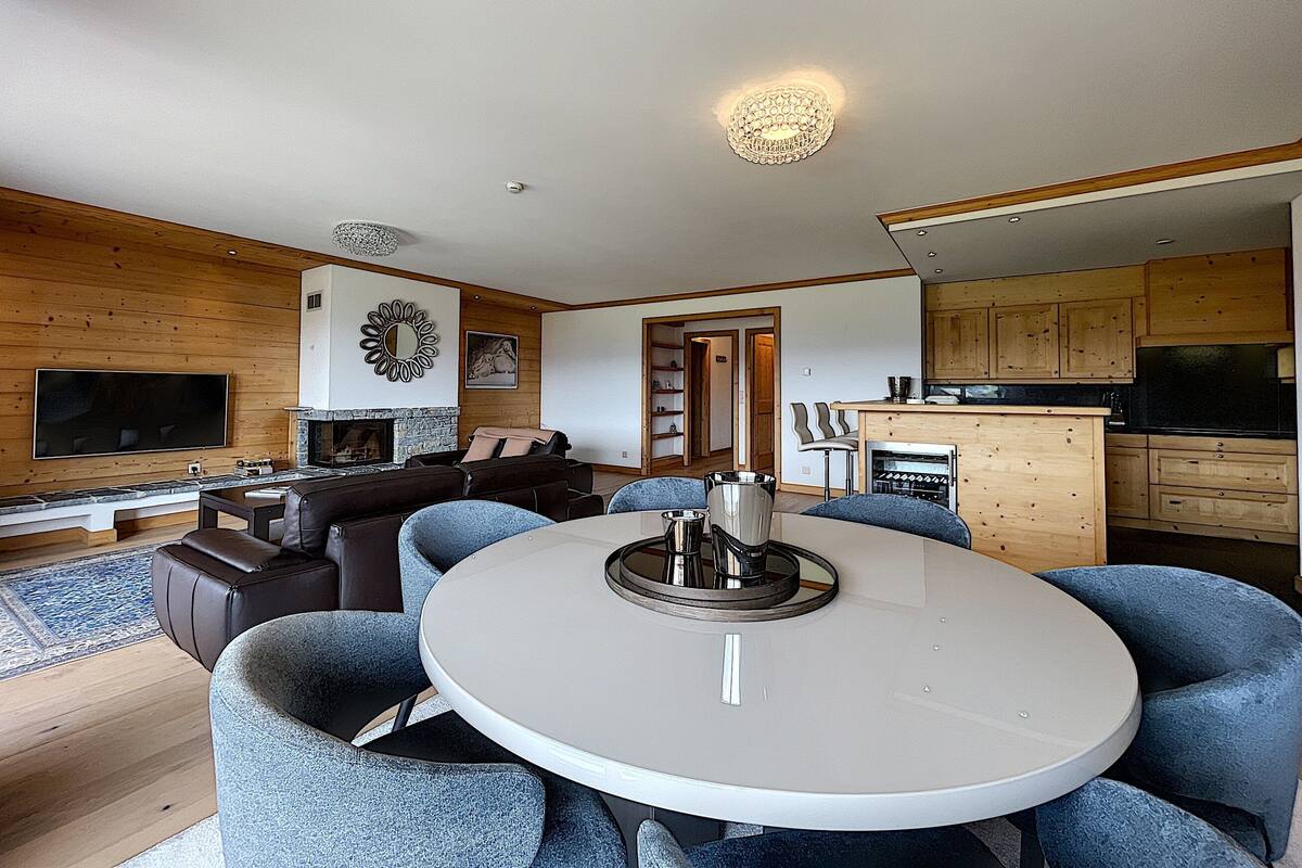 A modern living room with a fireplace, a dining table with chairs, a kitchen area, and wooden paneling on the walls.