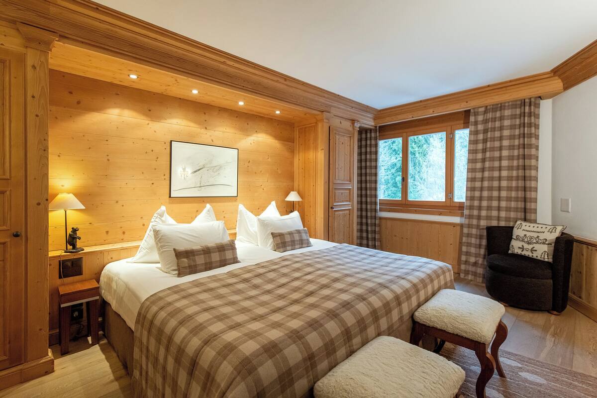 A cozy wooden bedroom with a large bed, two bedside tables, a chair, and a window with curtains.