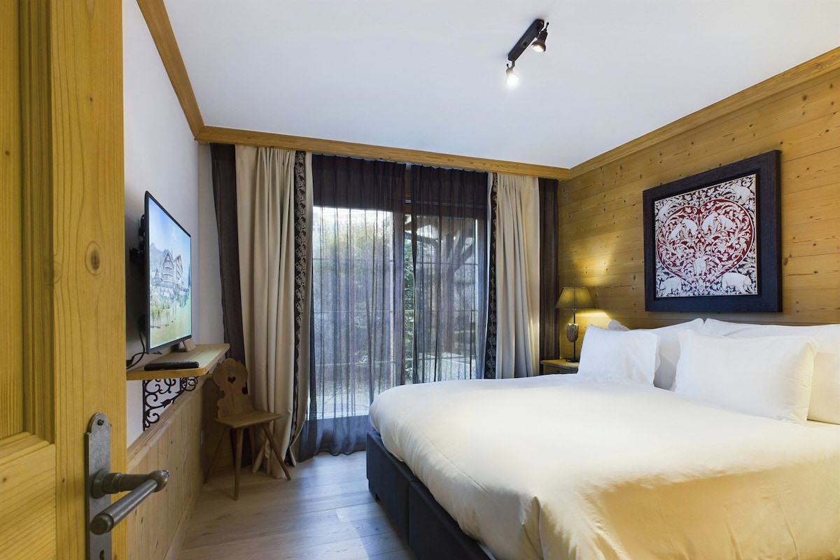 A hotel room with a large bed, a television on a wooden shelf, a nightstand with a lamp, and a window with curtains.