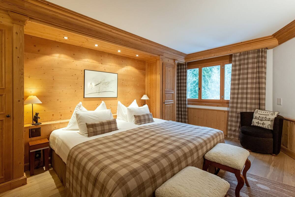 A wooden-paneled bedroom with a large bed, two bedside lamps, a chair, and a window with curtains.