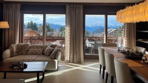 A spacious living room with a sofa, coffee table, and dining area, large windows with a mountain view, and modern lighting fixtures.