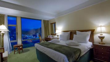 Quarto de hotel com uma cama grande, duas luminárias de cabeceira, uma mesa de cabeceira de madeira, uma mesinha e vista para a cidade através de uma janela ampla.