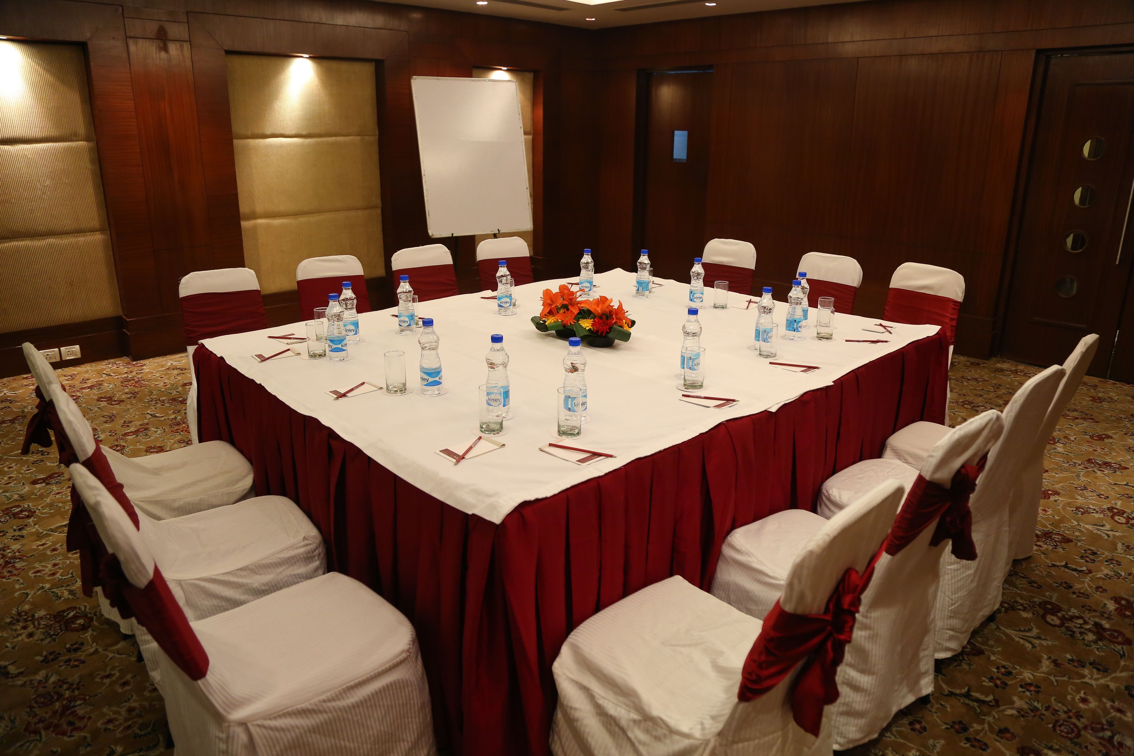 Une salle de conférence avec une grande table, des chaises blanches ornées de rubans rouges, des bouteilles d’eau et une composition florale en guise de centre de table.