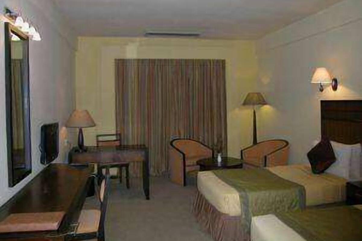 Quarto de hotel com cama, duas mesas de cabeceira, escrivaninha, cadeira, abajur e uma janela grande com cortinas.