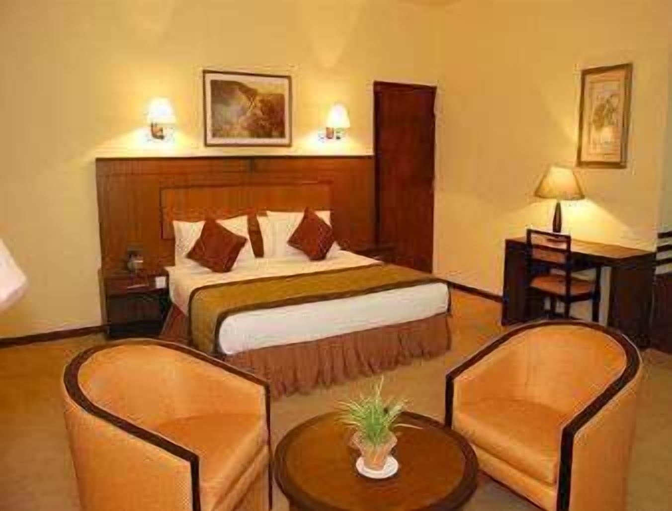 Une chambre d’hôtel comprenant un lit, deux fauteuils, une petite table avec une plante, un bureau avec une lampe et deux lampes fixées au mur.