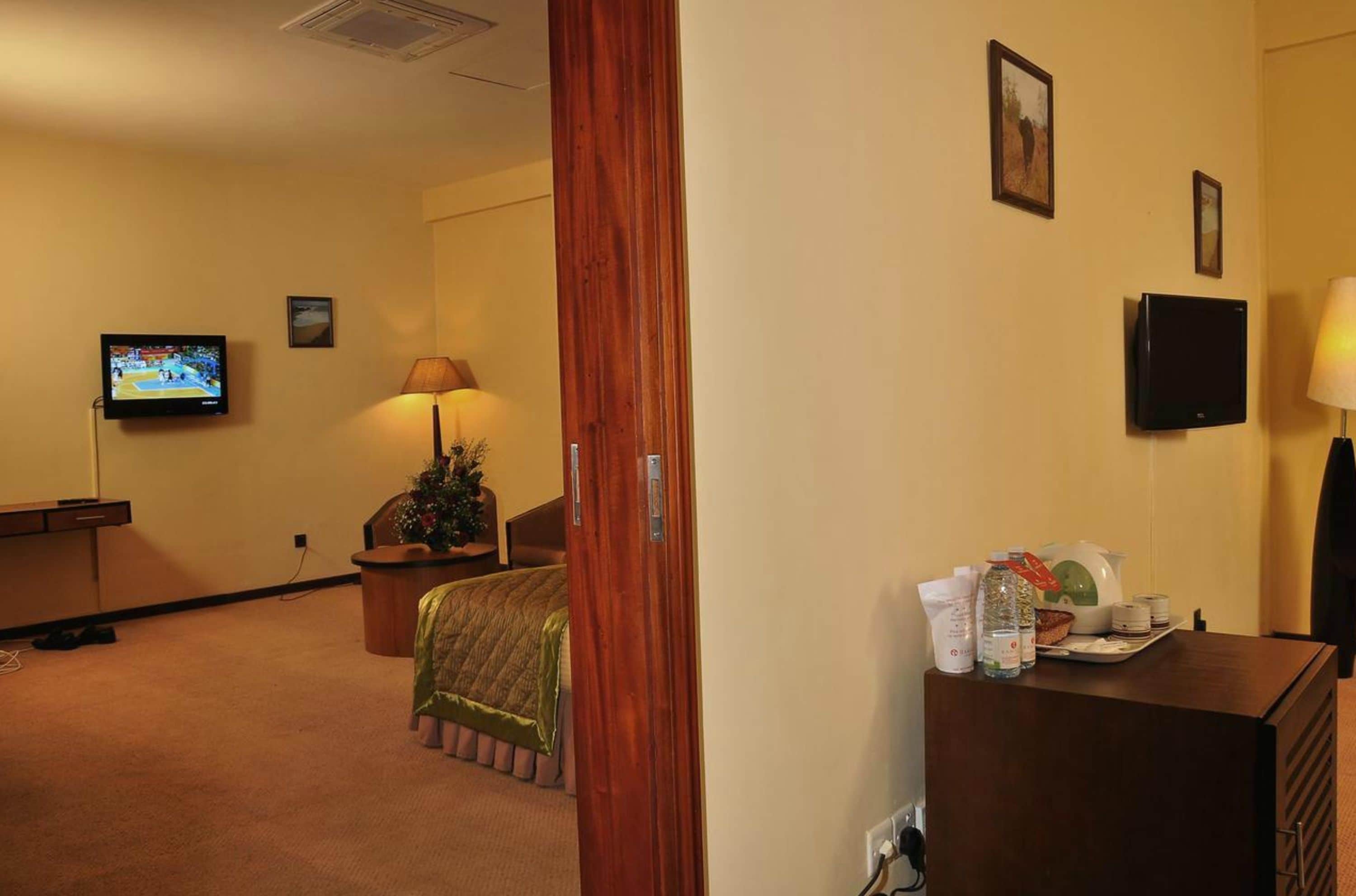 Habitación de hotel con cama, escritorio, televisor, lámpara y una mesita de noche con artículos de aseo.
