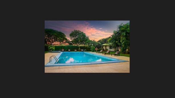 A rectangular swimming pool with a clear blue water surface, surrounded by a paved deck. There are lounge chairs and a small structure with a roof, possibly a changing room or a small building, in the background. - Ramada by Wyndham Katunayake Colombo International Airport (Seeduwa - Katunayake)