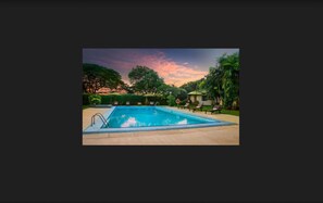 Una piscina rectangular con superficie de agua azul transparente, rodeada por una terraza pavimentada. Hay sillas de descanso y una estructura pequeña con techo al fondo, posiblemente un vestuario o un pequeño edificio.