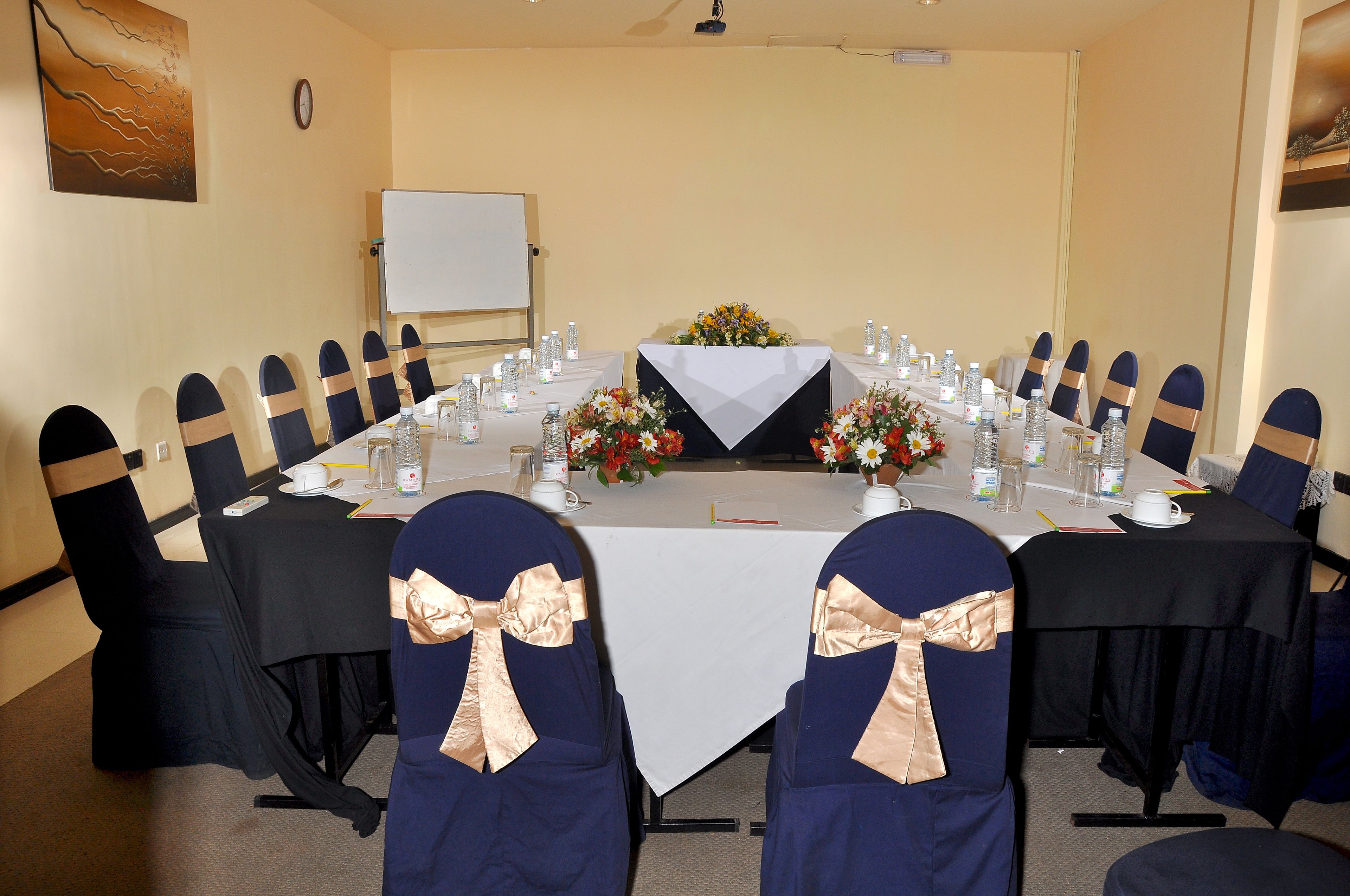 Une salle de conférence avec une longue table, des chaises décorées de nœuds, des bouteilles d’eau et des arrangements floraux en guise de centre de table.