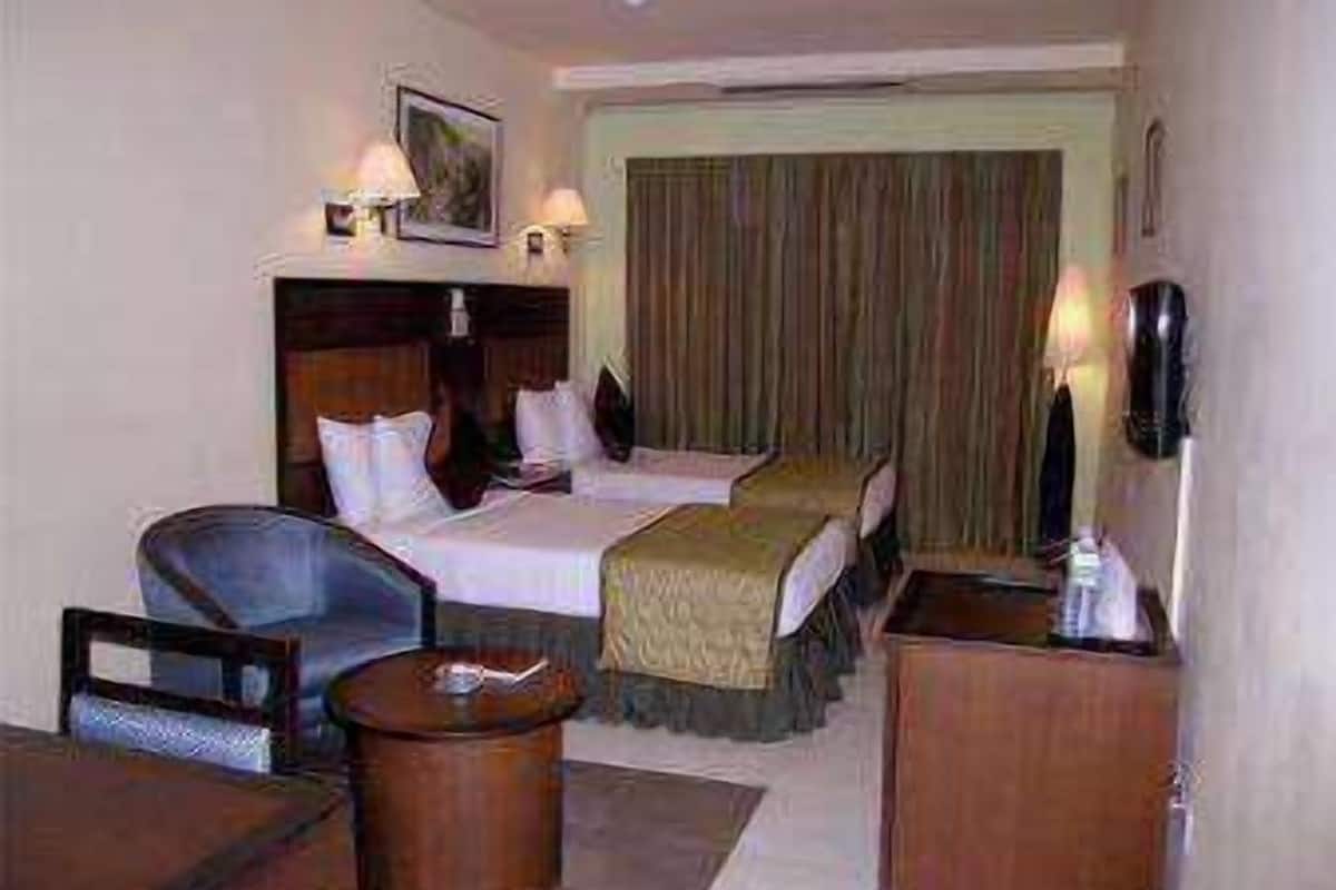 Quarto de hotel com duas camas, uma escrivaninha, uma cadeira, uma mesinha e uma mesa de cabeceira.