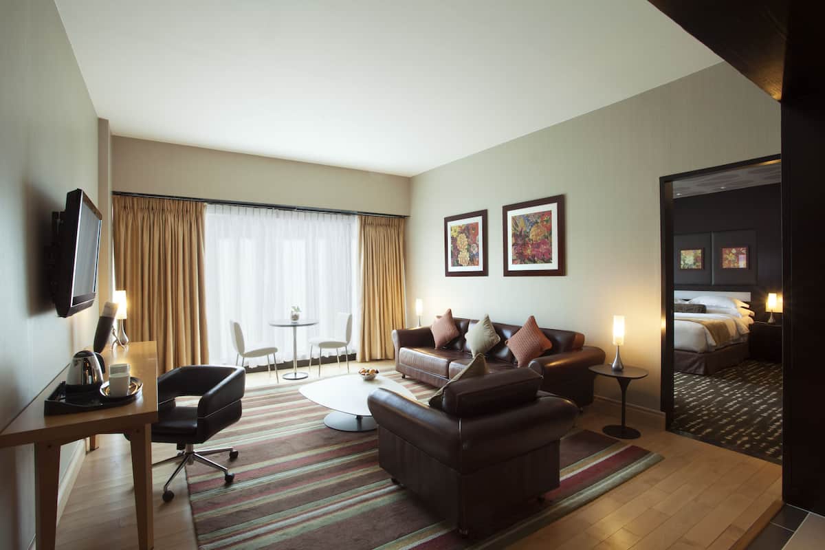A hotel room with a sofa, armchair, coffee table, and a TV on a stand. There is a small table with a lamp and a chair. The room has a large window with curtains and a door leading to a bedroom.