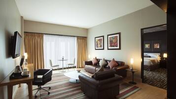 Une chambre d’hôtel avec un canapé, un fauteuil, une table basse et un téléviseur posé sur un meuble. On y trouve également une petite table avec une lampe et une chaise. La chambre est dotée d’une grande fenêtre avec des rideaux et d’une porte qui donne sur une chambre à coucher.