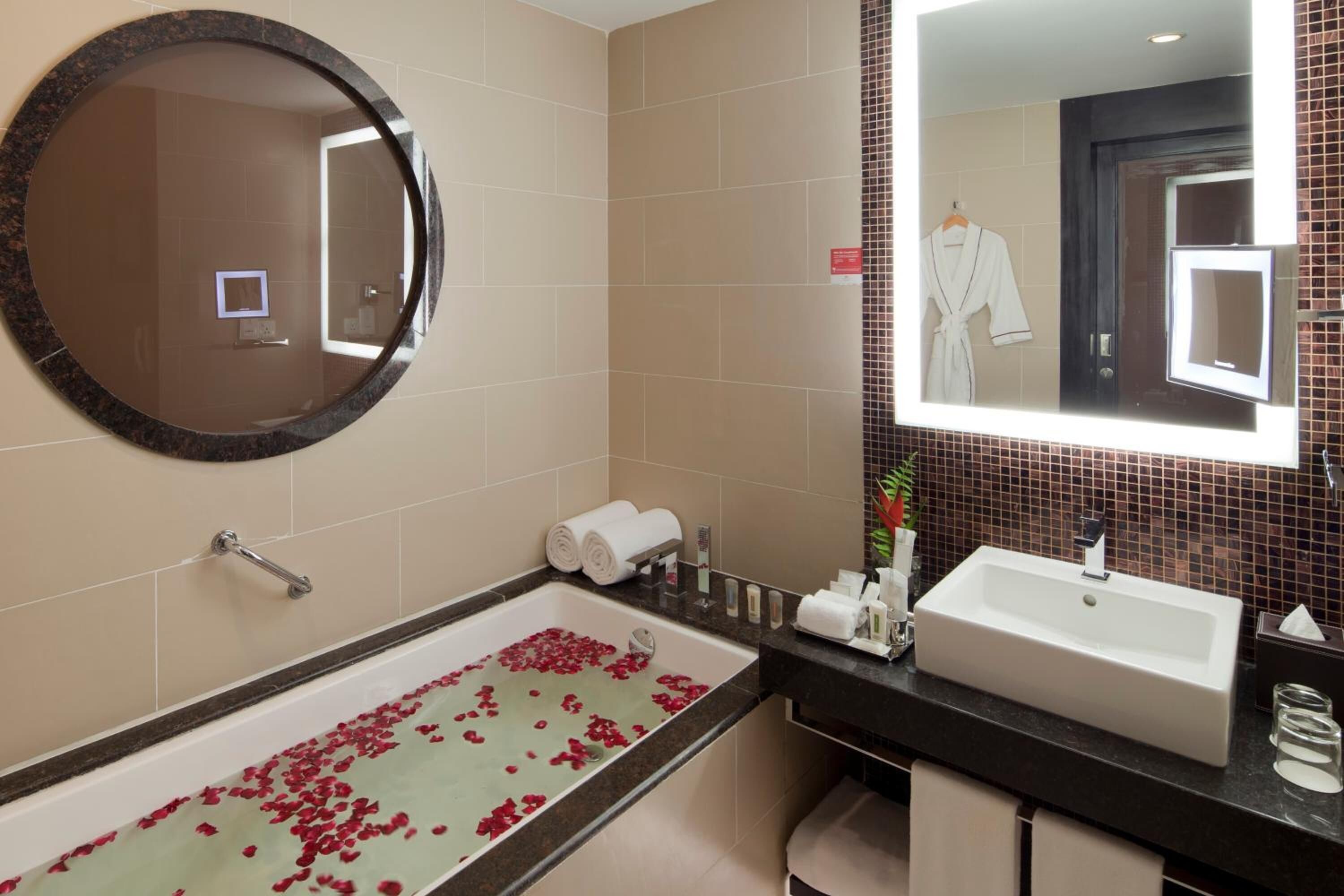 Un baño con una bañera llena de pétalos de rosa, un espejo redondo, un lavamanos y un perchero para toallas.