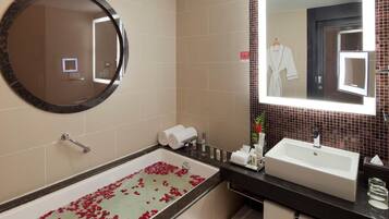 Un baño con una bañera llena de pétalos de rosa, un espejo redondo, un lavamanos y un perchero para toallas.