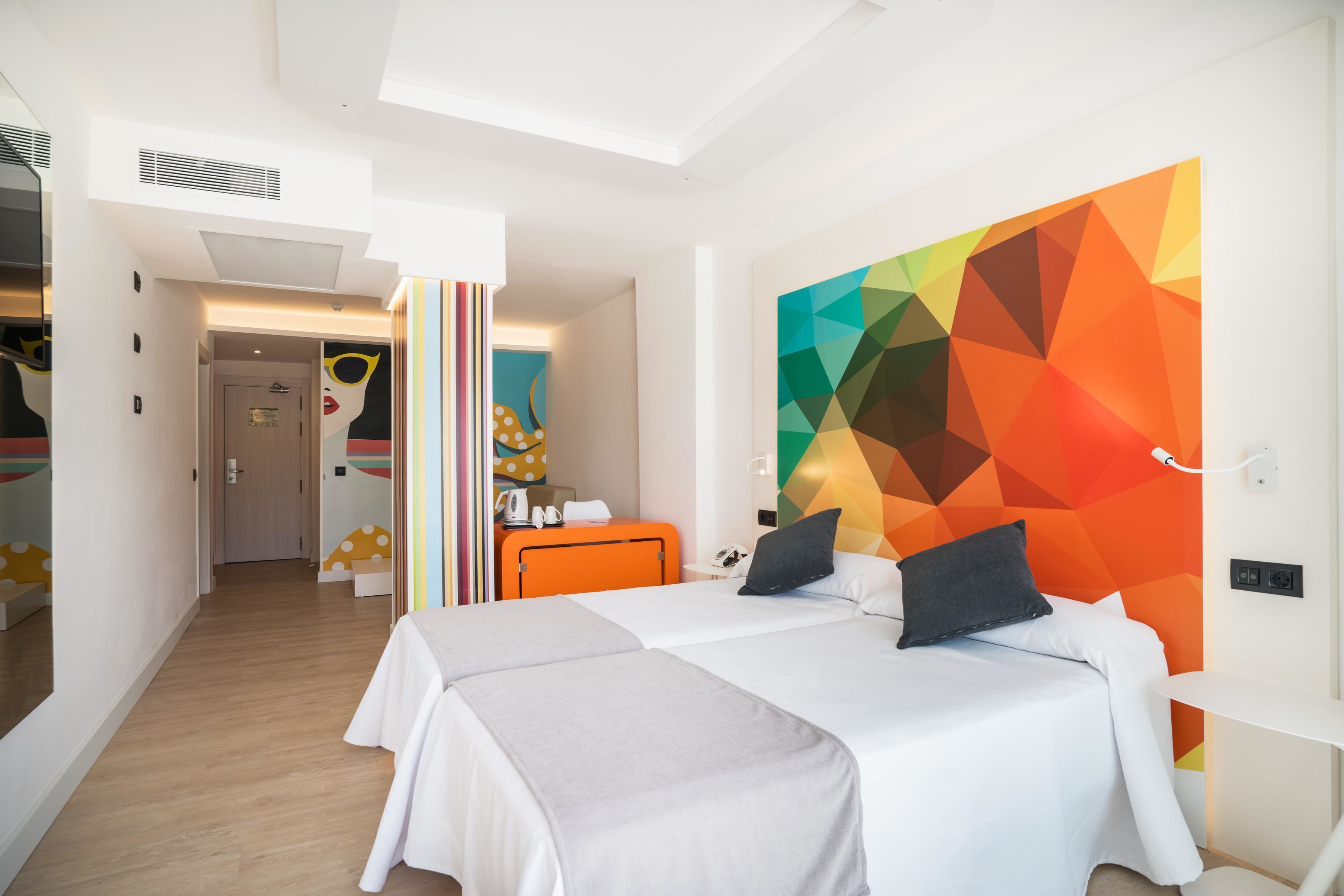 Una habitación de hotel moderna con dos camas, un colorido cuadro abstracto geométrico y un espacio luminoso y bien iluminado.