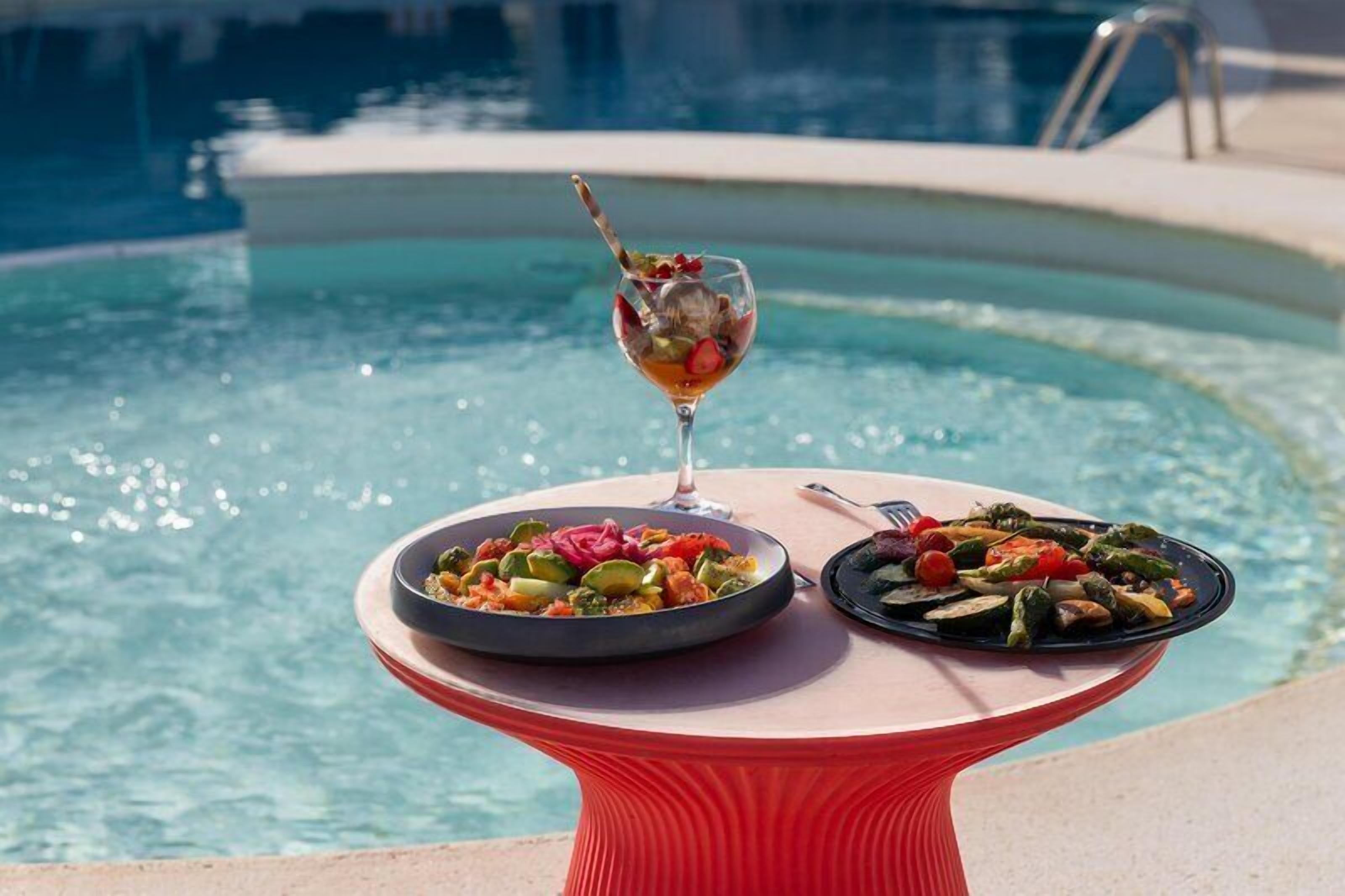 Ein Tisch mit zwei Tellern Essen und einem Getränk am Pool.