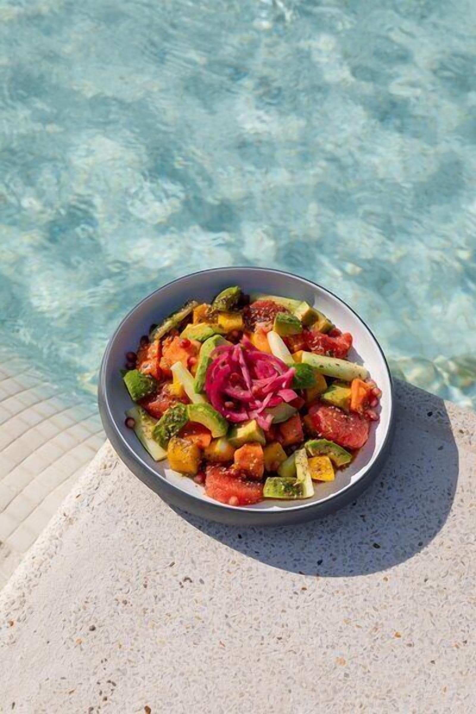 Un tazón de ensalada con diversos vegetales y un adorno rosa, sobre una superficie cerca de una piscina.