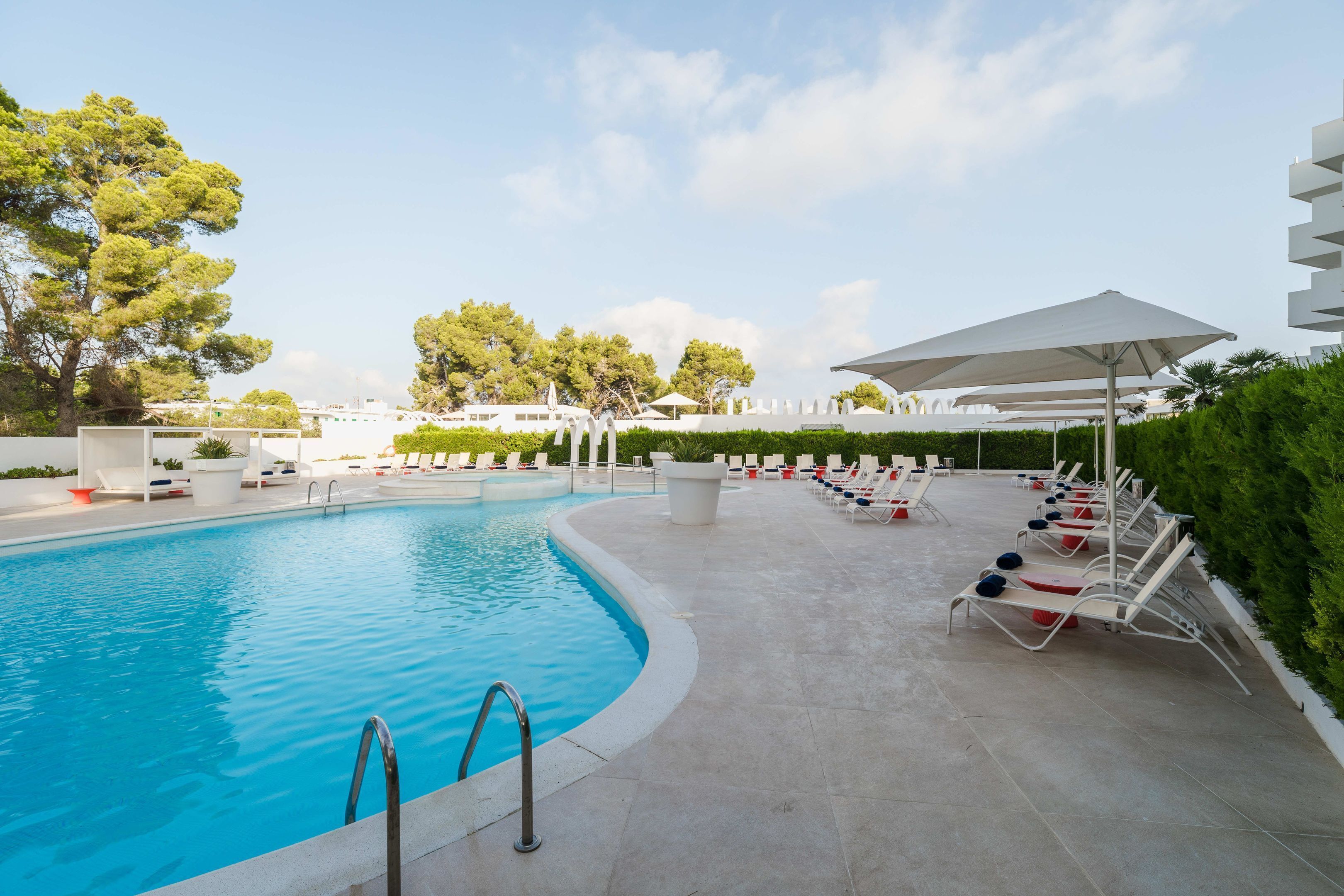 A swimming pool with lounge chairs and umbrellas, surrounded by greenery and white structures.