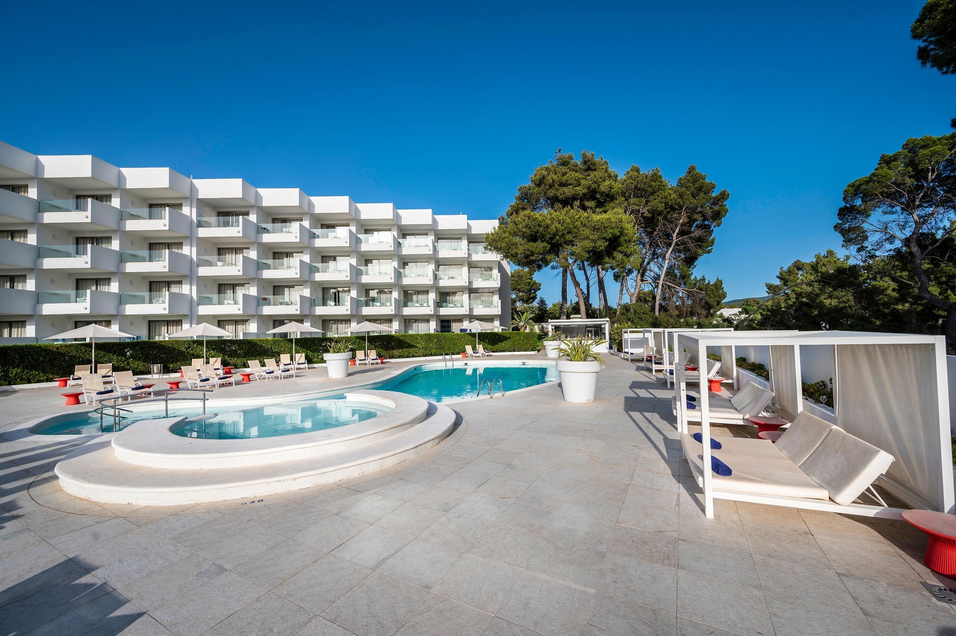 Un área de piscina con sillones de descanso, una piscina circular y un edificio con balcones al fondo.