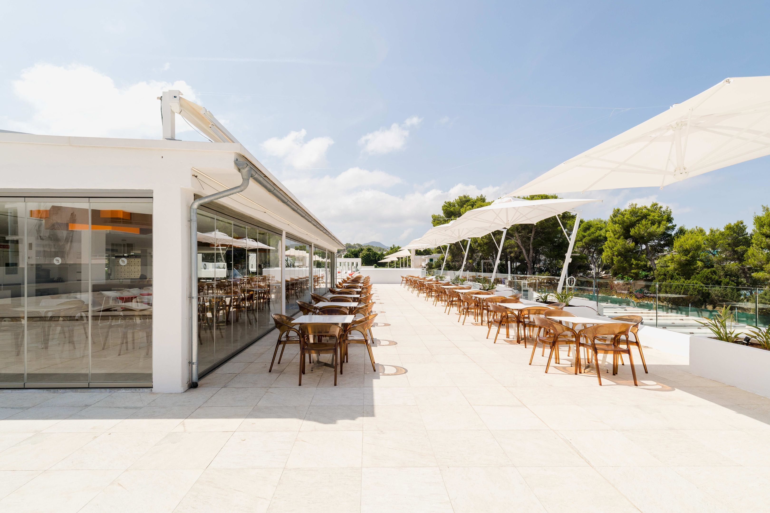 Area pranzo all'aperto con tavoli e sedie, grandi ombrelloni e un edificio in vetro adiacente alla zona salotto.