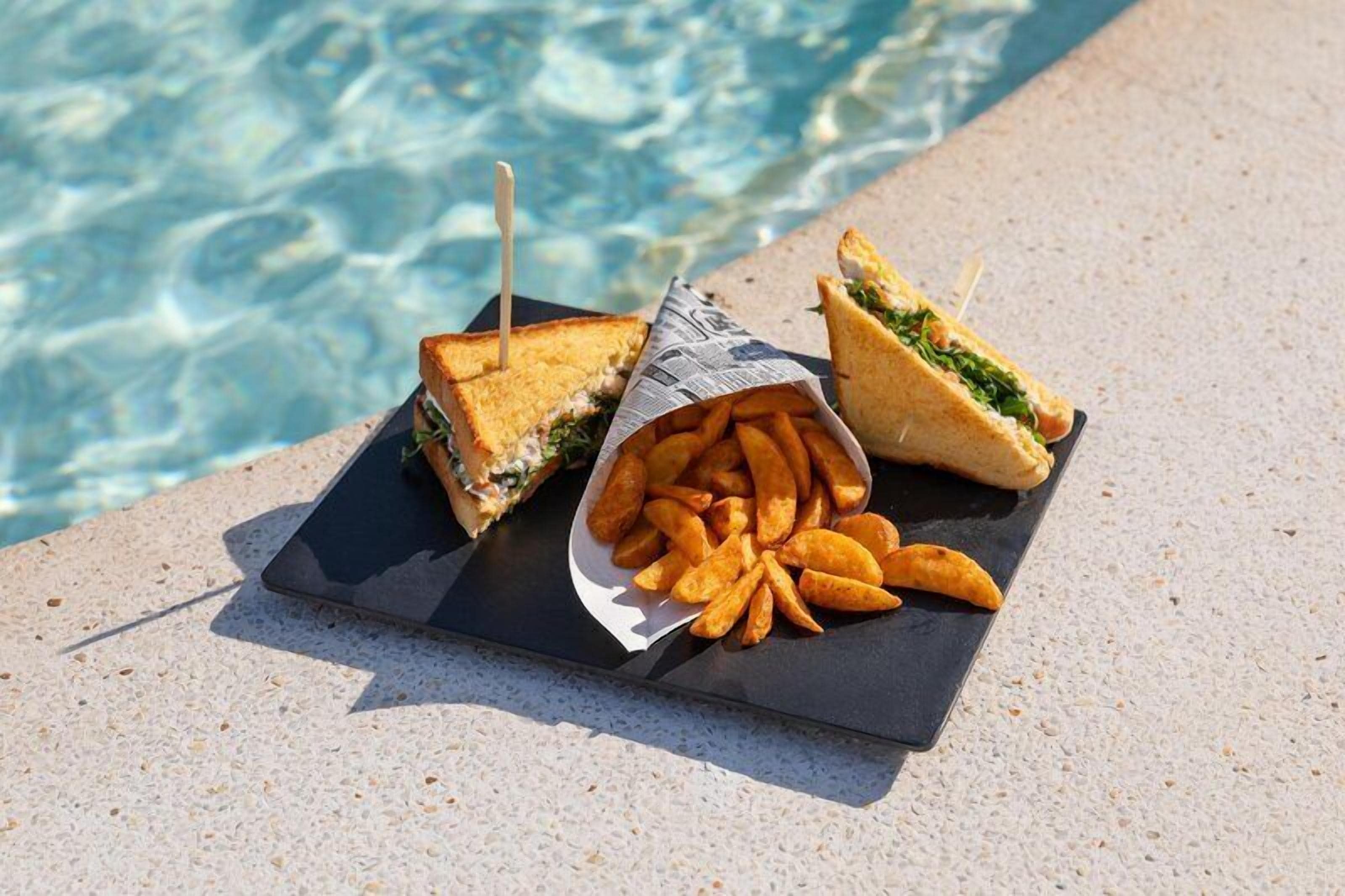 A poolside setting with a sandwich and fries on a black slate board.