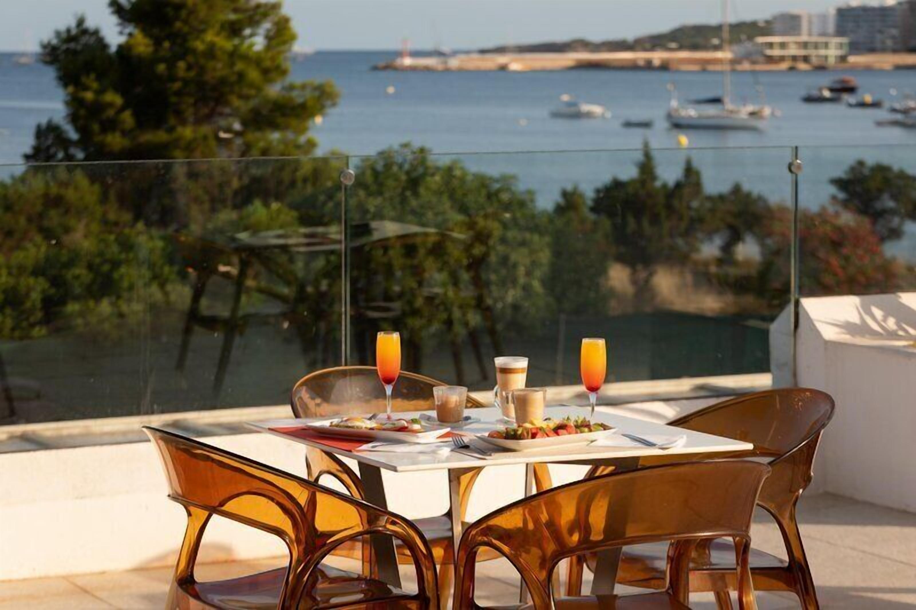 Une table dressée avec des aliments pour le petit-déjeuner, des chaises transparentes et une vue sur la mer et les bateaux.