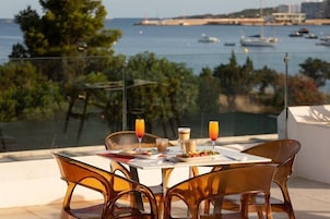 Una mesa con alimentos para el desayuno, sillas transparentes y vista al mar y embarcaciones.