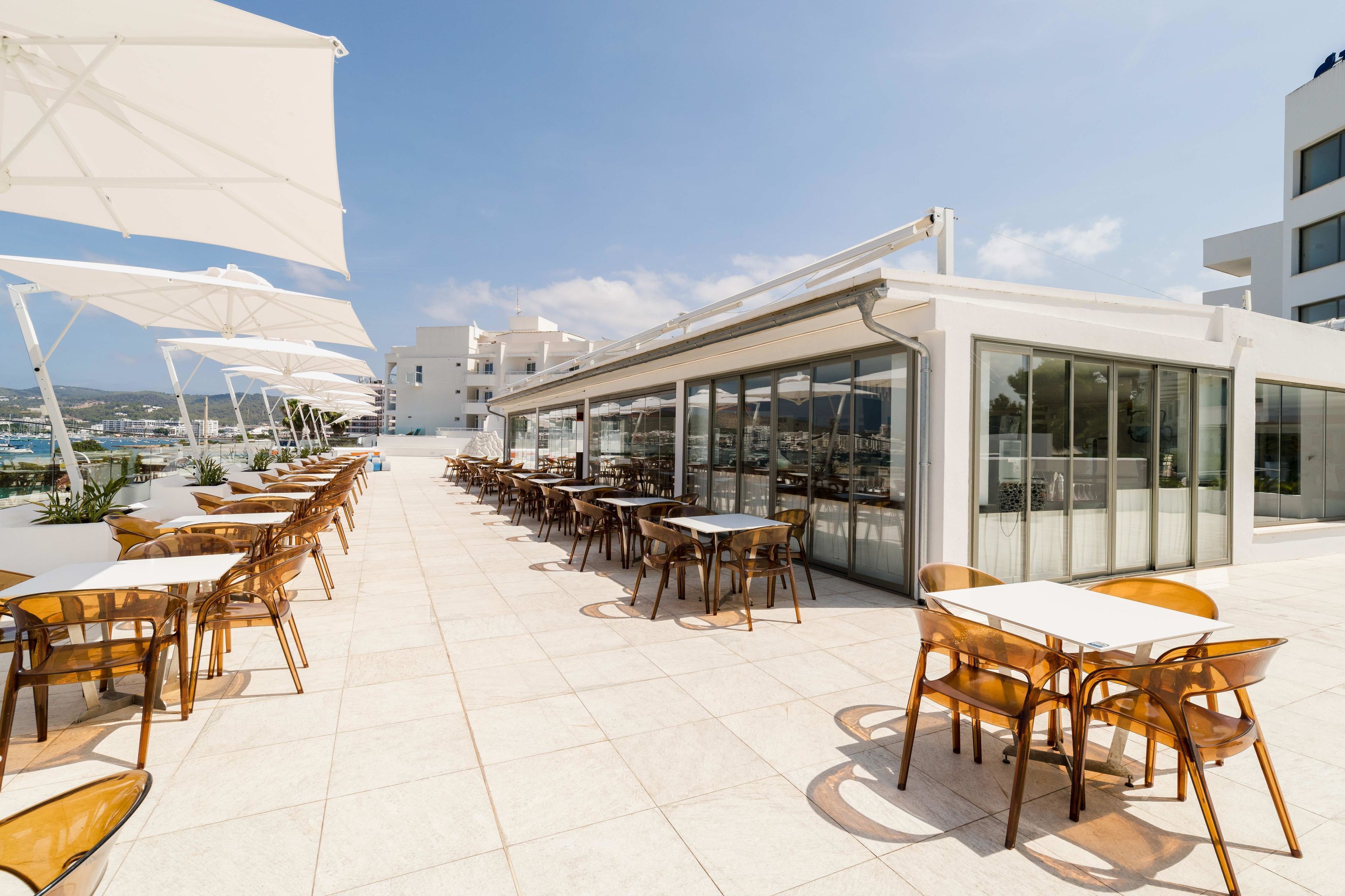 Una terraza en la azotea con mesas al aire libre, un bar con paredes de cristal y vistas panorámicas de la ciudad.