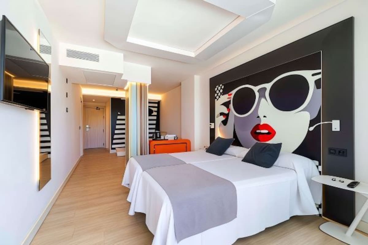 A modern hotel room with a large bed, a TV mounted on the wall, and a unique artwork featuring sunglasses and lips.